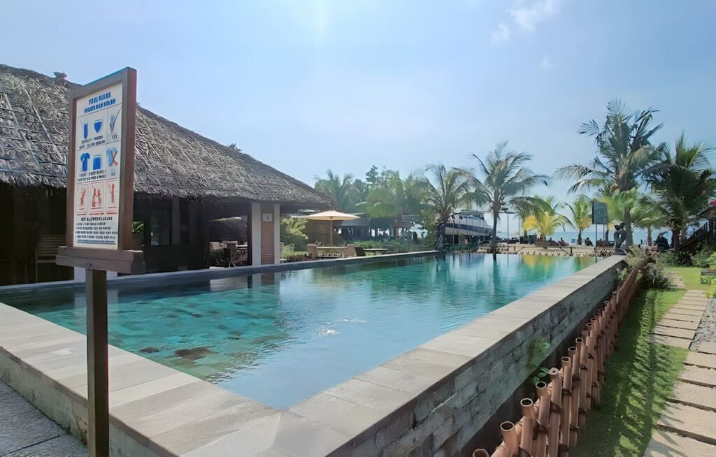 Kolam renang dengan air jernih di resort pantai, dikelilingi pepohonan dan tanda peraturan kolam.