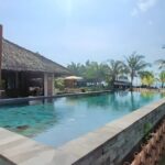 Kolam renang dengan air jernih di resort pantai, dikelilingi pepohonan dan tanda peraturan kolam.